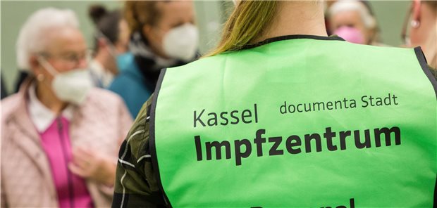 COVID-19 in Hessen: Ab Oktober übernehmen Haus- und Betriebsärzte das Impfen komplett Blick auf den Rücken einer Mitarbeiterin des Impfzentrums Kassel, verschwommen vor ihr Passanten unterschiedlichen Alters mit Masken.