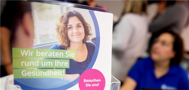 Ein Flyer für einen Gesundheitskiosk ist im Vordergrund zu sehen. Er trägt die Aufschrift: "Wir beraten Sie rund um Ihre Gesundheit" und "Besuchen Sie uns!". Im Hintergrund ist unscharf eine Frau im dunkelblauen Polohemd zu erkennen.