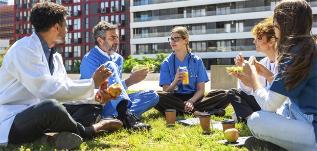 mehrere Ärzte sitzen im Kreis auf einer Wiese vor einer Klinikfassade