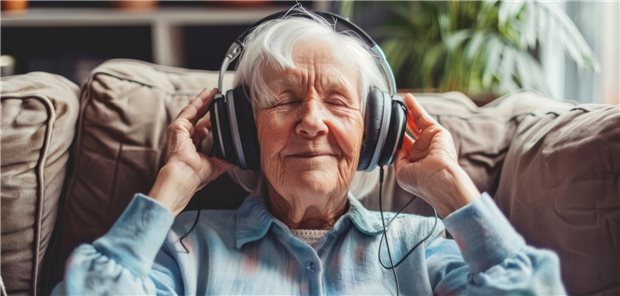 Eine ältere Frau hat dicke Kopfhörer auf und hört mit einem Lächeln Musik. 