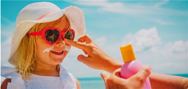 So werden Kinder richtig vor der Sonne geschützt! Mutter cremt Kind mit Sonnencreme ein – und auch ein Hut als Schutz ist gut.