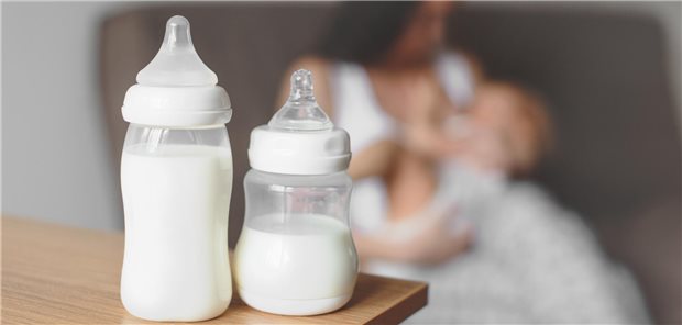Zwei Flaschen gefüllt mit Milch stehen im Vordergrund auf einem Tisch, dahinter stillt eine Frau ihr Baby.