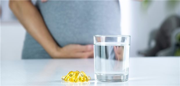 Eine Schwangere Frau im Hintergrund, im Vordergrund ein Glas Wasser und Kapseln.