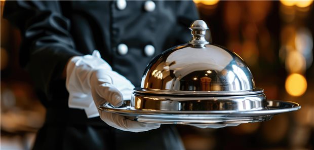 Ein Kellner mit weißen Handschuhen serviert ein Essen und unter einer silberner Tafelhaube.