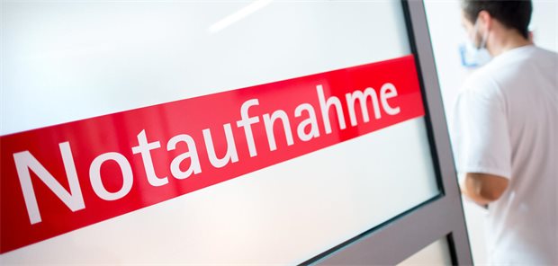  Ein Krankenpfleger steht in einem Krankenhaus neben einer Glastür mit der Aufschrift „Notaufnahme“. 