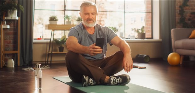 Ein älterer Herr sitzt im Schneidersitz auf einer Yoga-Matte und blickt auf sein Smartphone.