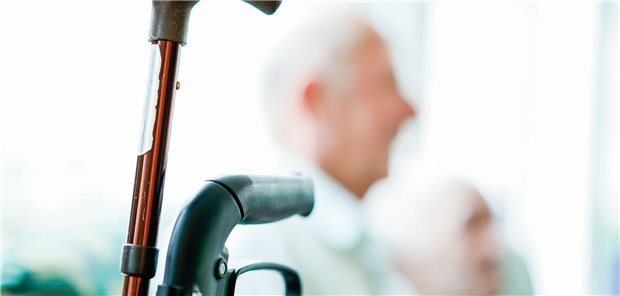 Krückstock an einem Rollator befestigt, im Hintergrund ein älterer Herr.