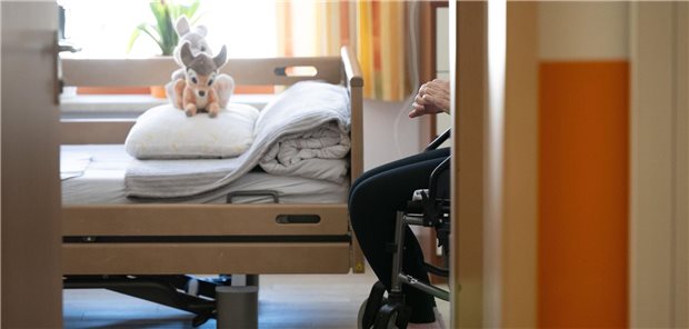 Blick in ein Zimmer in einem Pflegeheim, dort steht ein Bett, daneben sitzt eine pflegebedürftiger Mensch.