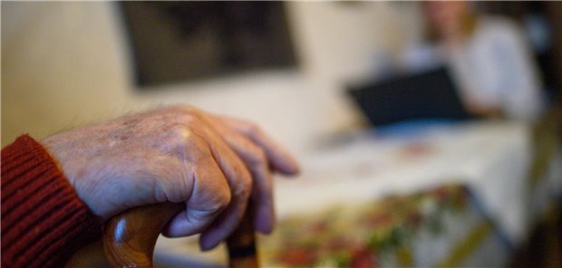 Die Hand einer pflegebedürftigen Seniorin an einem Gehstock; im Hintergrund einer Fachkraft mit Laptop.