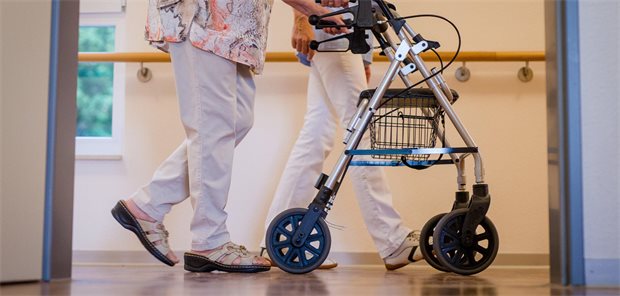 Eine Pflegekraft geht in einem Pflegeheim mit einer älteren Frau über einen Korridor. Die Frau schiebt einen Rollator.