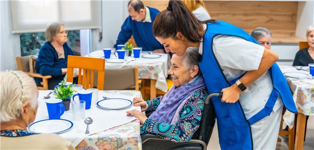 Eine Pflegekraft hilft einer älteren Frau beim Heranrücken an einen Esstisch.