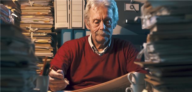 Älterer Mann sitzt in einem dunklen Raum umgeben von einem Stapel Akten.