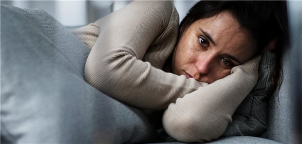 Eine Frau mit Depressionen liegt in embryonaler Haltung auf einer Couch und starrt ins Nichts.