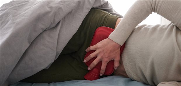 Eine Frau liegt im Bett und hält sich den Unterleib. Sie hat eine Wärmflasche auf dem Bauch.