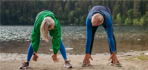 Eine Frau und ein Mann stehen in Sportklamotten vor einem See und versuchen mit den Händen den Boden zu berühren.