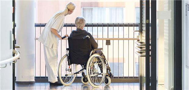 Ältere Patientin im Rollstuhl blickt nach draußen, neben ihr und ihr zugeneigt eine Pflegerin.