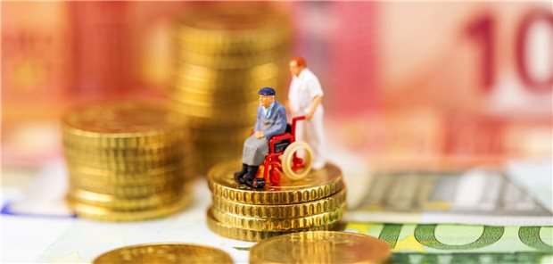 Miniaturfiguren: Ein Mann im Rollstuhl auf einer Geldmünze, hinter ihm eine Pflegekraft im mittleren Alter.