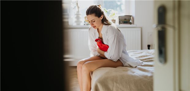 Das sind die aktuellen medikamentösen Ansätze bei Endometriose Eine Frau sitzt auf dem Bett und hält sich eine Wärmflasche auf den Bauch.