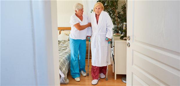 Blick ins Zimmer: Eine Pflegerin hilft einer älteren Dame auf Krücken.