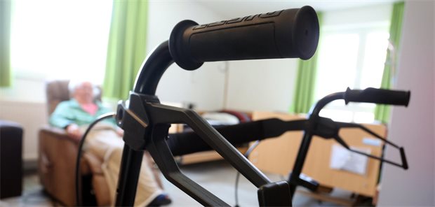Eine pflegebedürftige Frau sitzt in einem Apartment hinter einem Rollator.