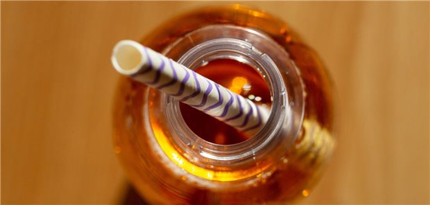 Eine geöffnete Flasche mit einem zuckerhaltigen Erfrischungsgetränk steht auf einem Tisch, in der Öffnung steckt ein Trinkhalm.