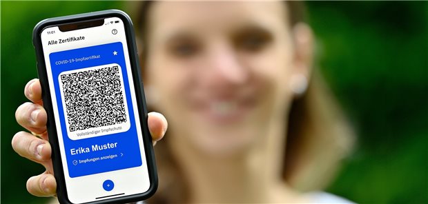 Corona-Daten erheben: „Wir stehen uns selbst im Weg“ Frau mit Smartphone in der Hand, auf dem die CovPass-App (für den digitalen Impfnachweis gegen SARS-CoV-2, COVID-19) zu sehen ist; die Dame ist unscharf im Hintergrund.