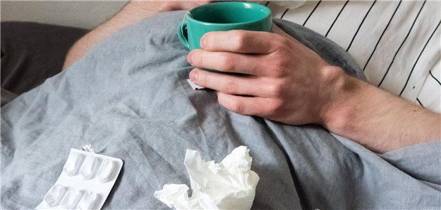 Ein Mann liegt mit Taschentüchern, Teetasse und Tabletten im Bett.