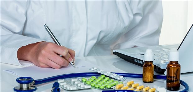 Person im Kittel schreibt etwas auf ein Papier, vor sich eine Vielzahl an Medikamenten, ein Stethoskop und ein Laptop.
