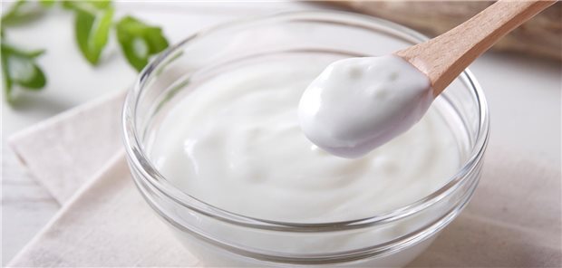Eine Schüssel Joghurth und ein Holzlöffel stehen auf einem Tisch mit hellem Tuch darauf.