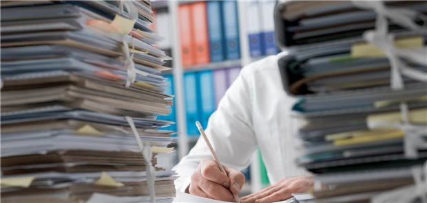 Mann sitzt am Schreibtisch. Vor ihm zwei Berge mit Dokumenten. 