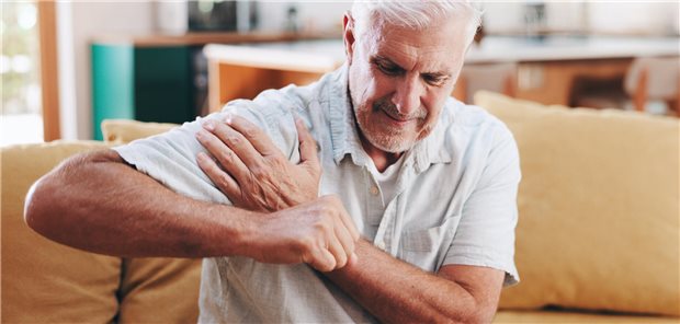 Ein Mann sitzt auf einem Sofa und legt die linke Hand auf die rechte Schulter, die schmerzt.
