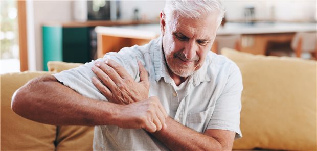 Ein Mann sitzt auf einem Sofa und legt die linke Hand auf die rechte Schulter, die schmerzt.
