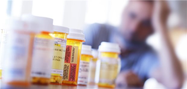 Viele Tablettendosen stehen auf einem Tisch. Im Hintergrund ist ein Mann zu sehen, der erschöpft seinen Kopf auf seiner Hand abstützt.