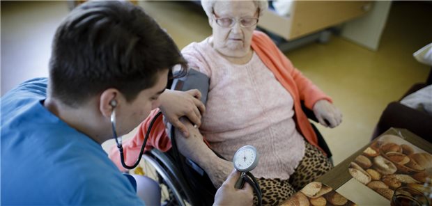 Eine männliche Pflegekraft misst Blutdruck bei einer älteren Frau im Rollstuhl.