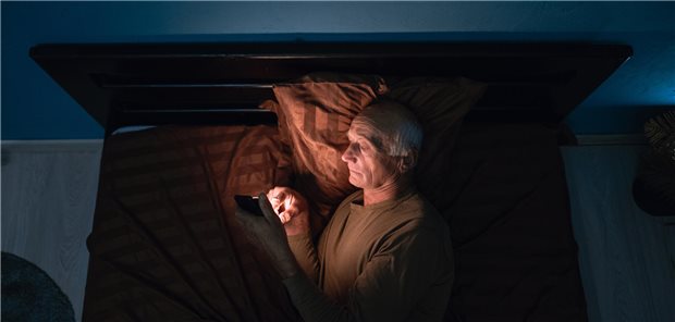 Wie sich schlechter Schlaf auf Schmerzen auswirkt Ein Mann liegt im Bett und schaut auf sein Handy.