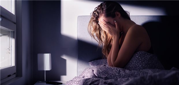Eine junge Frau sitzt im Bett und hält sich die Hände vors Gesicht.