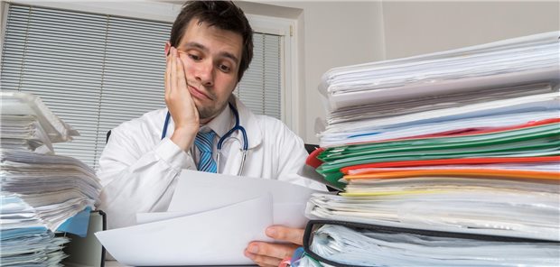 Studie: Mehr Personal durch gestiegene Bürokratie im Gesundheitswesen Ein Arzt schaut genervt auf Blätter, während dicke Aktenordnet um ihm herum liegen.