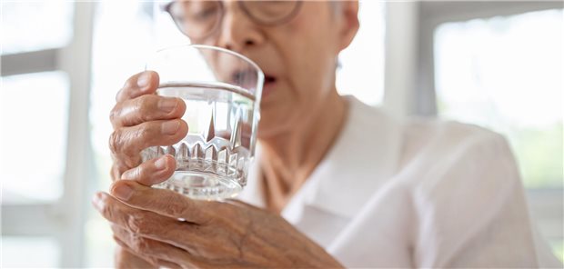 ältere asiatische Frau trinkt aus einem Wasserglas und stützt sich den Arm dabei