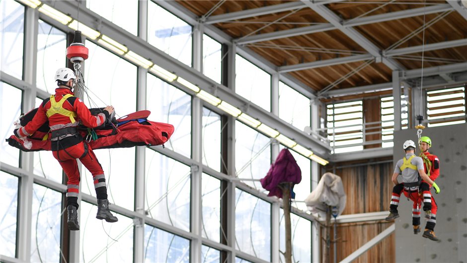 Zwei Teams von Notärzten sind gerade in der Luft – links wird ein Notarzt mit einem Bergesack hoch zur Hubschrauberzelle gewindet, rechts üben die Notärzte die Rettung mit Rettungsdreieck. Dabei handelt es sich um eine Art Tuch mit Karabinerhaken, das der Notarzt um die zu rettende Person zu einem Sitz faltet.